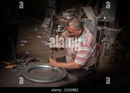 Ein Handwerker in Alexandria, Ägypten, der in einer Werkstatt arbeitet und eine Metallplatte sorgfältig von Hand formt, umgeben von traditionellen Werkzeugen und Materialien. Stockfoto