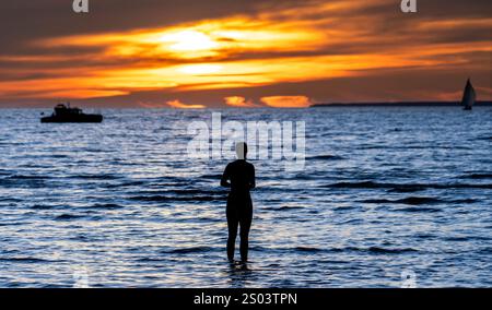 Eine Silhouette einer Person, die bei Sonnenuntergang im flachen Wasser steht, mit einem leuchtenden orange-gelben Himmel, der sich auf der Meeresoberfläche spiegelt. Ein Boot ist sichtbar Stockfoto