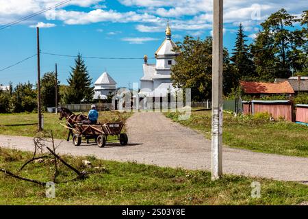 Ein Mann fährt mit einer Pferdekutsche eine Feldstraße hinunter. Die Szene ist friedlich und ruhig, mit einem kleinen Dorf im Hintergrund. Der Mann trägt sich Stockfoto
