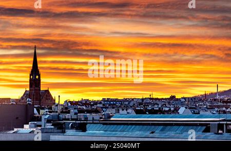 Ein atemberaubender Sonnenuntergang über einer Skyline der Stadt mit Dächern und einem markanten Kirchturm. Der Himmel ist voller leuchtender Orange- und Gelbtöne, Kreati Stockfoto