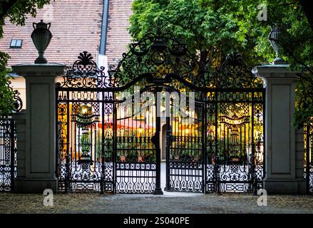 Ein wunderschön verziertes schmiedeeisernes Tor, das teilweise geöffnet ist, bietet einen Blick auf einen lebendigen Garten oder Innenhof dahinter. Die Szene wird von üppigem Grün eingerahmt Stockfoto