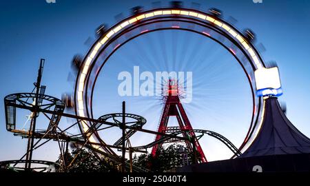 Eine lebhafte Szene im Vergnügungspark mit einem großen Riesenrad, das vor einem Dämmerungshimmel beleuchtet wird. Das Rad bewegt sich und erzeugt einen Unschärfeeffekt Stockfoto
