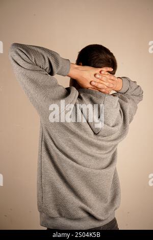 Ein Mann in einem Hoodie schließt seine Hände hinter den Kopf und dehnt sich in einer entspannten Haltung. Der isolierte Hintergrund unterstreicht sein beiläufiges, friedliches Verhalten. Stockfoto