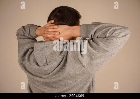 Ein Mann in einem Hoodie schließt seine Hände hinter den Kopf und dehnt sich in einer entspannten Haltung. Der isolierte Hintergrund unterstreicht sein beiläufiges, friedliches Verhalten. Stockfoto