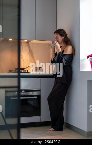 Frau im schwarzen Seidenschlafanzug steht in der modernen Küche und trinkt aus einem Glas. Sie lehnt sich an eine Wand in der Nähe von eleganten Schränken und Geräten. Stockfoto