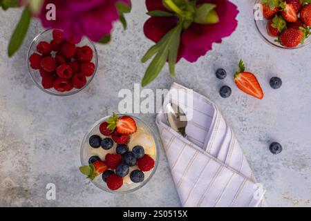 Blick von oben auf ein Sommerdessert mit Vanilleeis, frischen Beeren in einer Glasschale neben einer weißen Serviette, Löffel und leuchtenden rosafarbenen Pfingstrosen Stockfoto