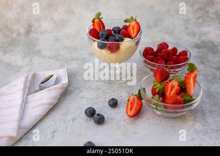 Erfrischendes Vanilleeis, serviert mit frischen Beeren in einer Glasschale, umgeben von beerengefüllten Gerichten auf einer strukturierten hellen Oberfläche Stockfoto