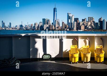 Einzigartige Perspektive auf Lower Manhattan & One World Trade Center, erfasst von der Fähre Liberty Island, eingerahmt von gelben Reinigungswagen und einem blauen Himmel. Stockfoto