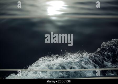 Eine Nahaufnahme von Wellen, die gegen die Seite eines Bootes brechen, wobei das schimmernde Sonnenlicht vom ruhigen Meer reflektiert wird Stockfoto