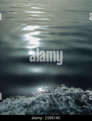 Eine Nahaufnahme von Wellen, die gegen die Seite eines Bootes brechen, wobei das schimmernde Sonnenlicht vom ruhigen Meer reflektiert wird Stockfoto