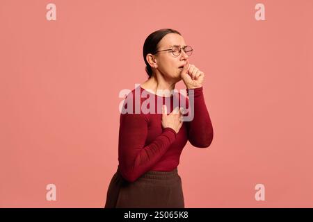 Frau in weinrot oben hustet in die Faust, andere Hand auf der Brust, Augen schielen leicht, stehend vor rosa Hintergrund. Stockfoto