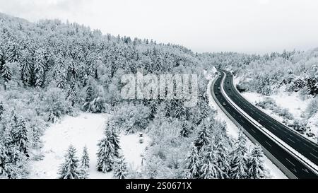 Ein atemberaubender Blick aus der Luft auf eine kurvenreiche Autobahn, die durch einen dichten, schneebedeckten Wald führt. Stockfoto