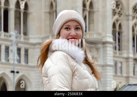 Eine schöne Frau im Wintermantel posiert vor einem historischen Gebäude und lächelt an einem kalten Tag in der Stadt Stockfoto