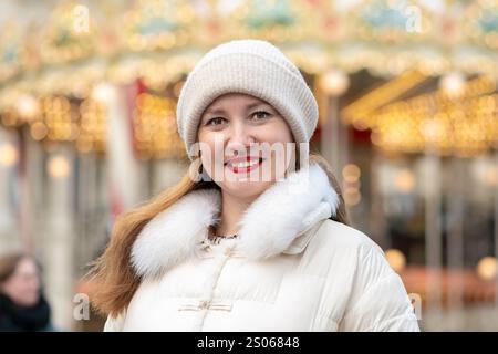 Eine 30-35-jährige Frau steht in einer stilvollen weißen Daunenjacke und Strickmütze auf einem geschäftigen Wintermarkt. Ihr selbstbewusstes Lächeln strahlt Wärme aus Stockfoto