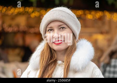 Eine 30-35-jährige Frau steht in einer stilvollen weißen Daunenjacke und Strickmütze auf einem geschäftigen Wintermarkt. Ihr selbstbewusstes Lächeln strahlt Wärme aus Stockfoto