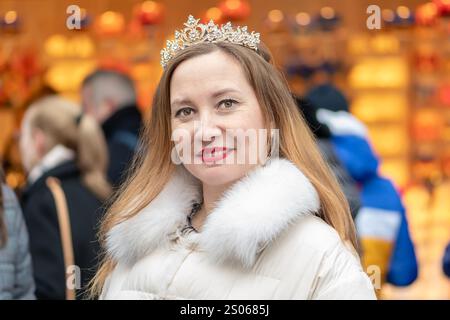 Eine Frau in Krone und Wintermantel lächelt während der Weihnachtszeit warm vor einem festlichen Stadtbild. Stockfoto