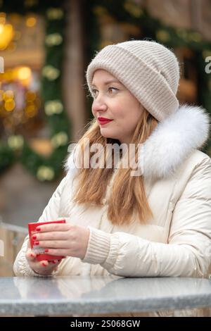 Eine Frau mit einem warmen Getränk, gekleidet in einem gemütlichen Wintermantel und Perlen, umgeben von funkelnden Lichtern und festlichen Dekorationen in urbaner Atmosphäre. Stockfoto
