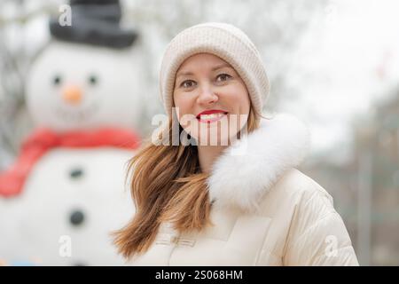 Winterporträt einer Frau, die vor einem Schneemann lächelt, der für den Urlaub dekoriert ist, draußen in einem verschneiten Park Stockfoto
