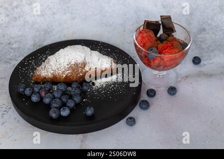 Dessertkomposition mit einem frischen Croissant, das mit Puderzucker bestäubt ist, und einer Glasschale mit Beerensorbet mit dunklen Schokoladenstückchen Stockfoto