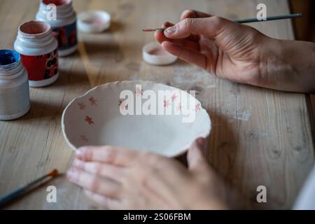 Handwerkliche Hände, die Muster auf handgefertigten Keramikplatten auf Holztisch malen. Kunstwerke der Töpferschule Stockfoto
