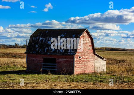 Eine rote Scheune sitzt auf einem Feld mit blauem Himmel im Hintergrund. Die Scheune ist alt und hat ein verwittertes Aussehen. Nostalgiekonzept und Verbindung zu Stockfoto