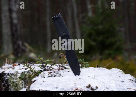 Ein schwarzes taktisches Messer mit fester Klinge steckt in einem Baumstamm in einem Winterwald. Stockfoto