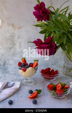 Elegante Dessertkomposition mit frischen Beeren und Eis in Glasschalen, begleitet von einem Bouquet von tiefen magentafarbenen Pfingstrosen in einer Vase Stockfoto