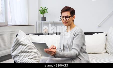 Reife Frau mit Brille, die bequem auf dem Sofa sitzt und mit einem Laptop die Geschenkoptionen auf einem Online-Marktplatz erkundet Stockfoto