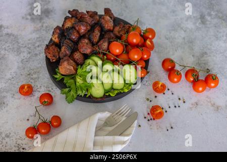 Köstliche gegrillte Fleischspieße, serviert mit frischen Kirschtomaten, Gurkenscheiben und Petersilie auf einem schwarzen Teller, ergänzt durch Messer und Gabel Stockfoto