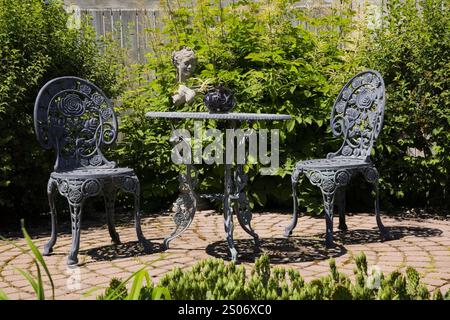 Graue gusseiserne Bistrotisch und Stühle auf der braunen Terrasse aus Pflasterstein im rustikalen Garten im Sommer. Stockfoto