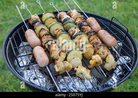 Nahaufnahme eines Barbecue-Grills mit Hühnerspießen, Zucchini und Würstchen, die über heißen Kohlen gekocht werden, umgeben von Kartoffeln in Folienfolie Stockfoto