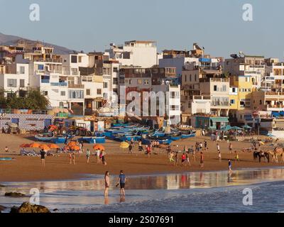 Paare im Wasser mit Menschen und blauen Fischerbooten an einem Sandstrand im Fischerdorf Taghazout, Marokko. Dezember 2024. Stockfoto
