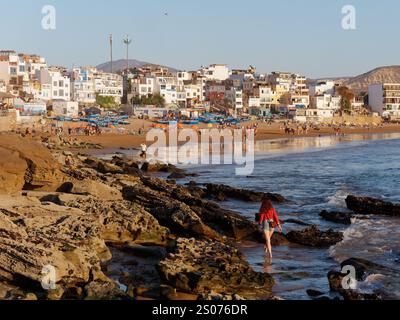 Sandstrand mit blauen Fischerbooten als Frau in Rot entlang der felsigen Küste im Fischerdorf Taghazout, Marokko. Dezember 2024. Stockfoto