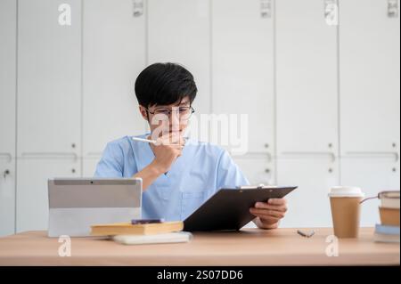 Ein kluger asiatischer männlicher Medizinstudent in Peelings, konzentriert sich auf die Überprüfung medizinischer Fälle auf einem Klemmbrett an seinem Schreibtisch in einer Bibliothek oder im Klassenzimmer. Medizin und Peo Stockfoto