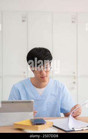 Ein kluger asiatischer männlicher Medizinstudent in Peelings, konzentriert sich auf die Überprüfung medizinischer Fälle auf einem Klemmbrett an seinem Schreibtisch in einer Bibliothek oder im Klassenzimmer. Medizin und Peo Stockfoto