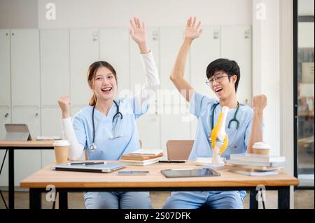 Zwei fröhliche asiatische männliche und weibliche Medizinstudenten in Peelings heben ihre Hände, um ihre Leistung zu feiern, während sie an einem Tisch und Hengst sitzen Stockfoto