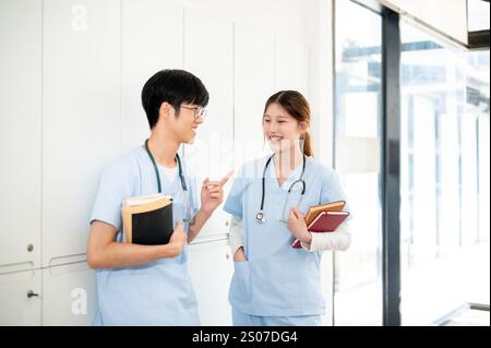 Zwei glückliche asiatische Ärztinnen oder Medizinstudenten tragen ihre Lehrbücher, während sie in der Umkleidekabine stehen und reden. Medizinstudium Stockfoto