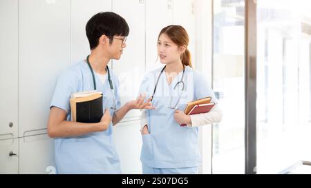 Zwei glückliche asiatische Ärztinnen oder Medizinstudenten tragen ihre Lehrbücher, während sie in der Umkleidekabine stehen und reden. Medizinstudium Stockfoto