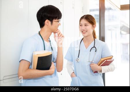 Zwei glückliche asiatische Ärztinnen oder Medizinstudenten tragen ihre Lehrbücher, während sie in der Umkleidekabine stehen und reden. Medizinstudium Stockfoto