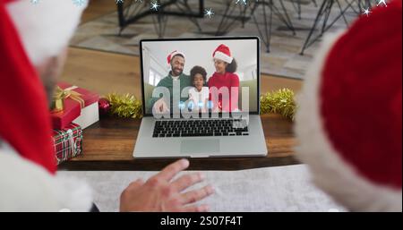 Bild von Schnee, der über lächelndes afroamerikanisches Paar in santa-Hüten fällt, auf Laptop-Image-Anruf Stockfoto