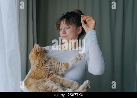 Die junge Frau hält eine Ingwerkatze, die bis zu ihrem Haar reicht, während sie sie in der Nähe eines Fensters mit transparenten Vorhängen justiert Stockfoto