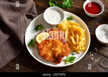 Köstliches paniertes Schnitzel mit Kartoffelfritten und Sauce auf weißem Teller auf Holztisch. Nahansicht Stockfoto