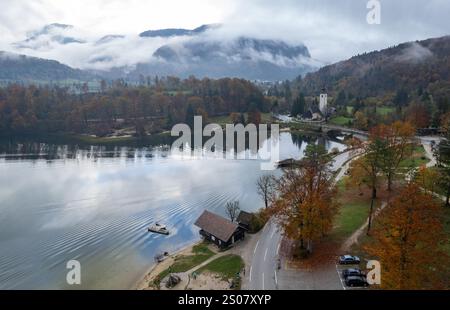 Die Drohnenszene des Motorbootes segelt auf dem Bohinj-See in der Nähe der Wallfahrtskirche St. Johannes der täufer, umgeben von bunten Herbstbäumen und Nebel Stockfoto