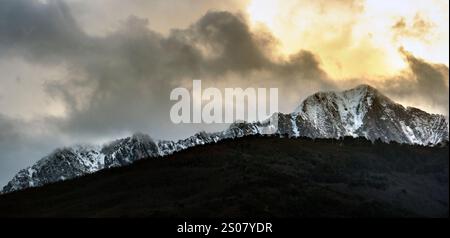 Eine dramatische Berglandschaft mit schneebedeckten Gipfeln unter bewölktem Himmel mit einem goldenen Farbton der untergehenden Sonne. Stockfoto