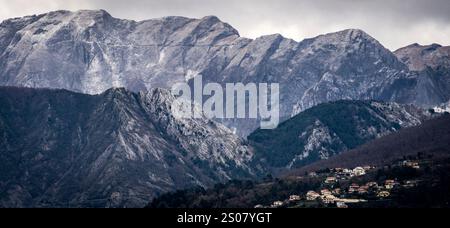 Eine dramatische Berglandschaft mit zerklüfteten Gipfeln und bewölktem Himmel. Der Vordergrund zeigt ein kleines Dorf, eingebettet zwischen den Hügeln, mit Bäumen und Ro Stockfoto