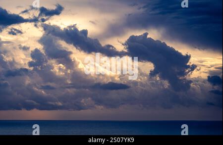 Ein dramatischer Himmel voller dunkler, wirbelnder Wolken über einem ruhigen Ozean. Die Wolken werden durch sanftes Licht beleuchtet, was einen Kontrast zu den tiefblauen s bildet Stockfoto