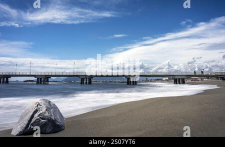 Eine ruhige Strandszene mit einem großen Felsen im Vordergrund, sanften Wellen am Ufer und einem langen Pier, der sich unter einem blauen Meer in den Ozean erstreckt Stockfoto