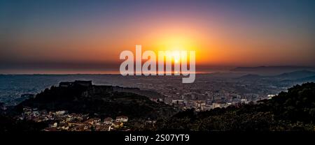 Ein atemberaubender Sonnenuntergang über einer Küstenstadt, während die Sonne unter dem Horizont eintaucht. Die Landschaft bietet Hügel und ein weitläufiges Stadtgebiet mit cas Stockfoto