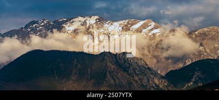 Ein atemberaubender Blick auf schneebedeckte Berge, die teilweise von Wolken bedeckt sind, mit rauem Gelände und dramatischer Beleuchtung. Stockfoto