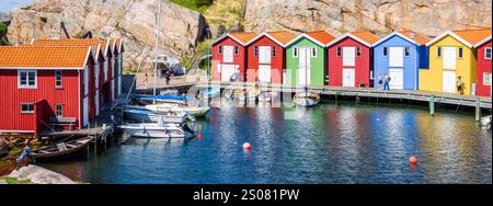 Panoramablick auf farbenfrohe hölzerne Bootshäuser, die sich an einem sonnigen Sommertag an der felsigen Klippe entlang des Meereskanals in Smögen, Schweden, aneinanderreihen. Stockfoto
